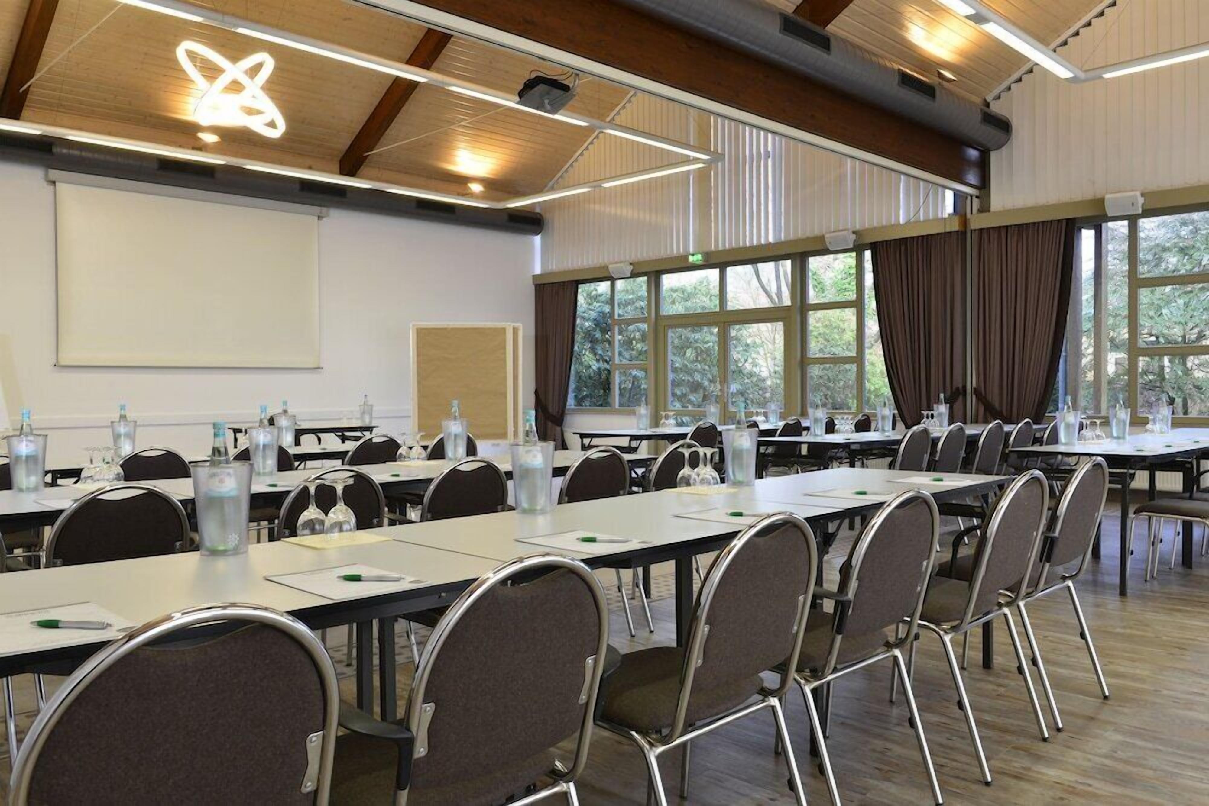 Un salón de conferencias con mesas largas, sillas, una pantalla de proyector y ventanas grandes con cortinas.