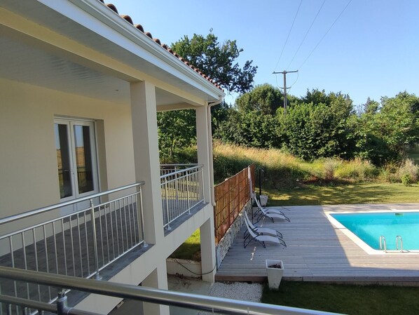 Zona junto a la piscina con terraza de madera, baranda metálica y vista a un paisaje verde.