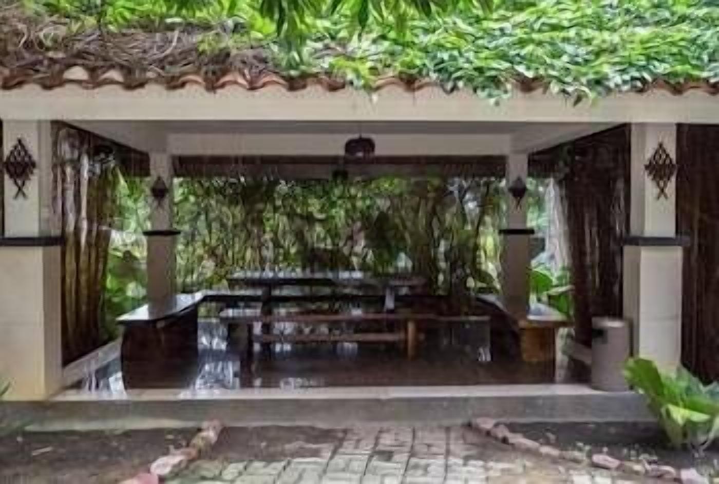 A covered outdoor seating area with a bench and a small table, surrounded by lush greenery and a tiled floor.