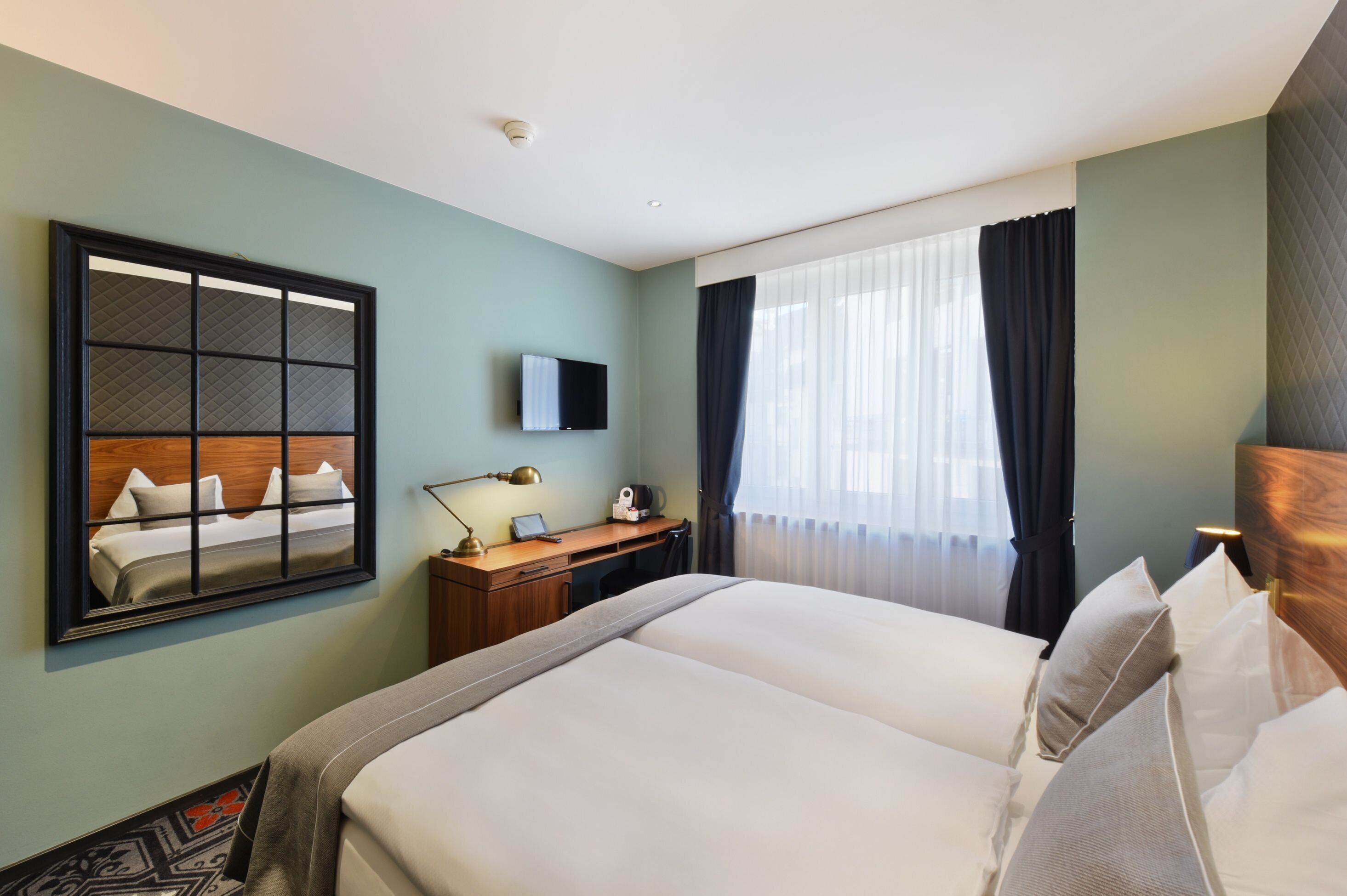 A hotel room with a large bed, a desk with a chair, a flat-screen TV, a window with curtains, and a wall-mounted mirror.
