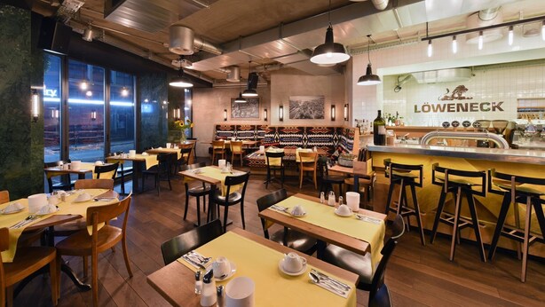 Un interior de restaurante con mesas y sillas de madera, una zona de bar y amplias ventanas que ofrecen vistas al exterior.
