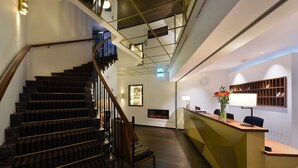Una escalera con barandilla de madera y peldaños negros que conduce a una zona de recepción con un mostrador, sillas y un estante en la pared.