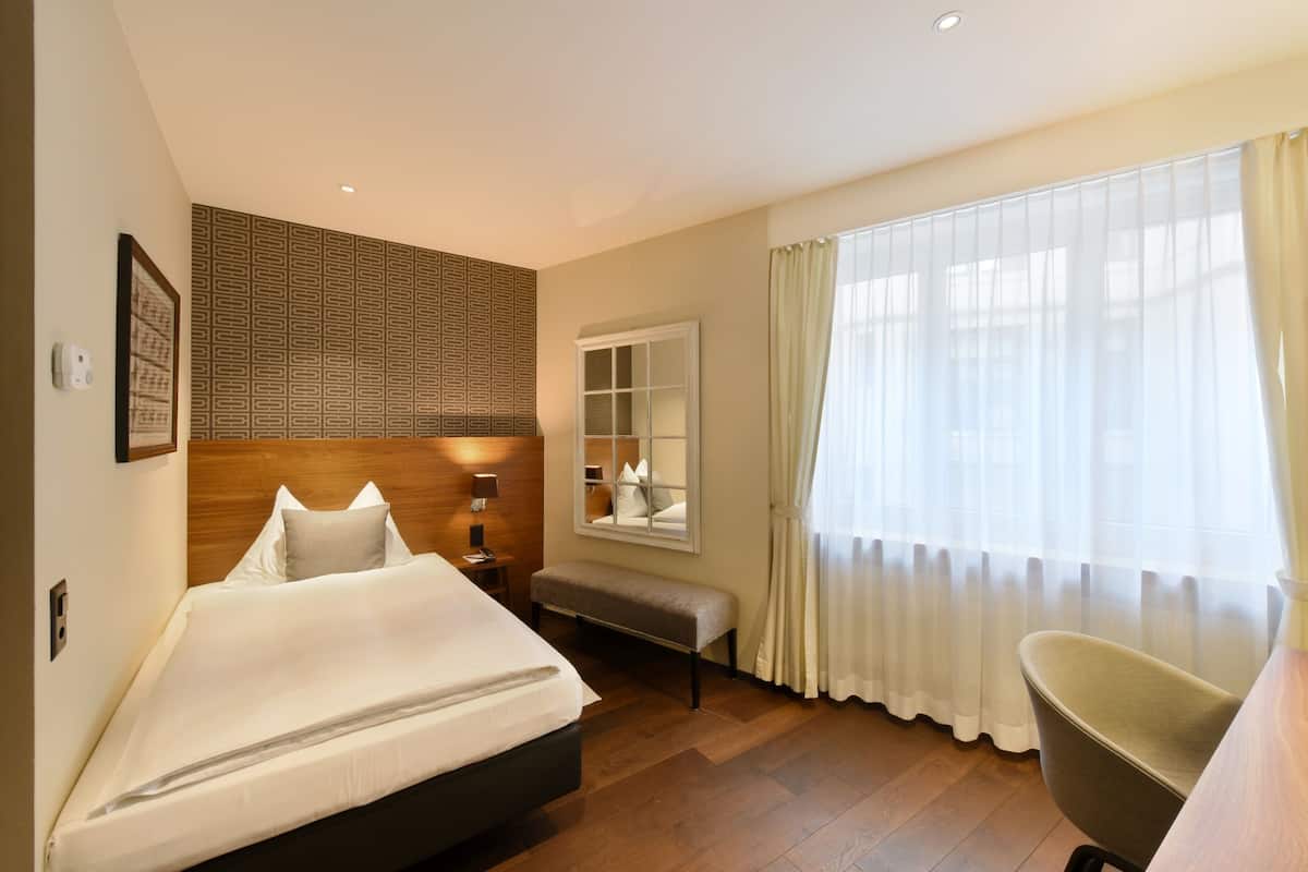 A hotel room with a bed, a bench, a chair, a bedside table, a mirror, and a window with curtains.