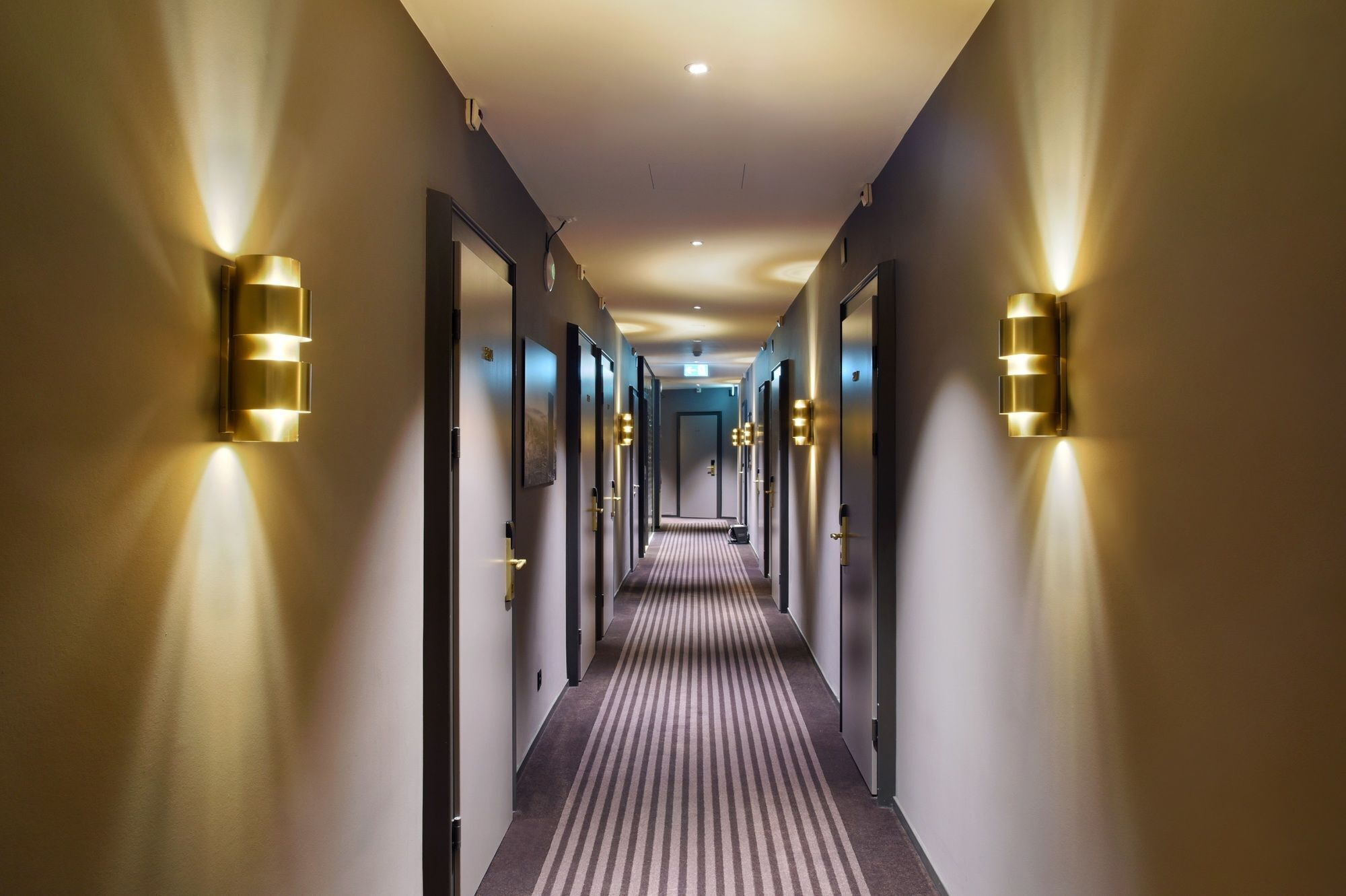A hotel hallway with a striped carpet, doors, and wall-mounted lighting fixtures.