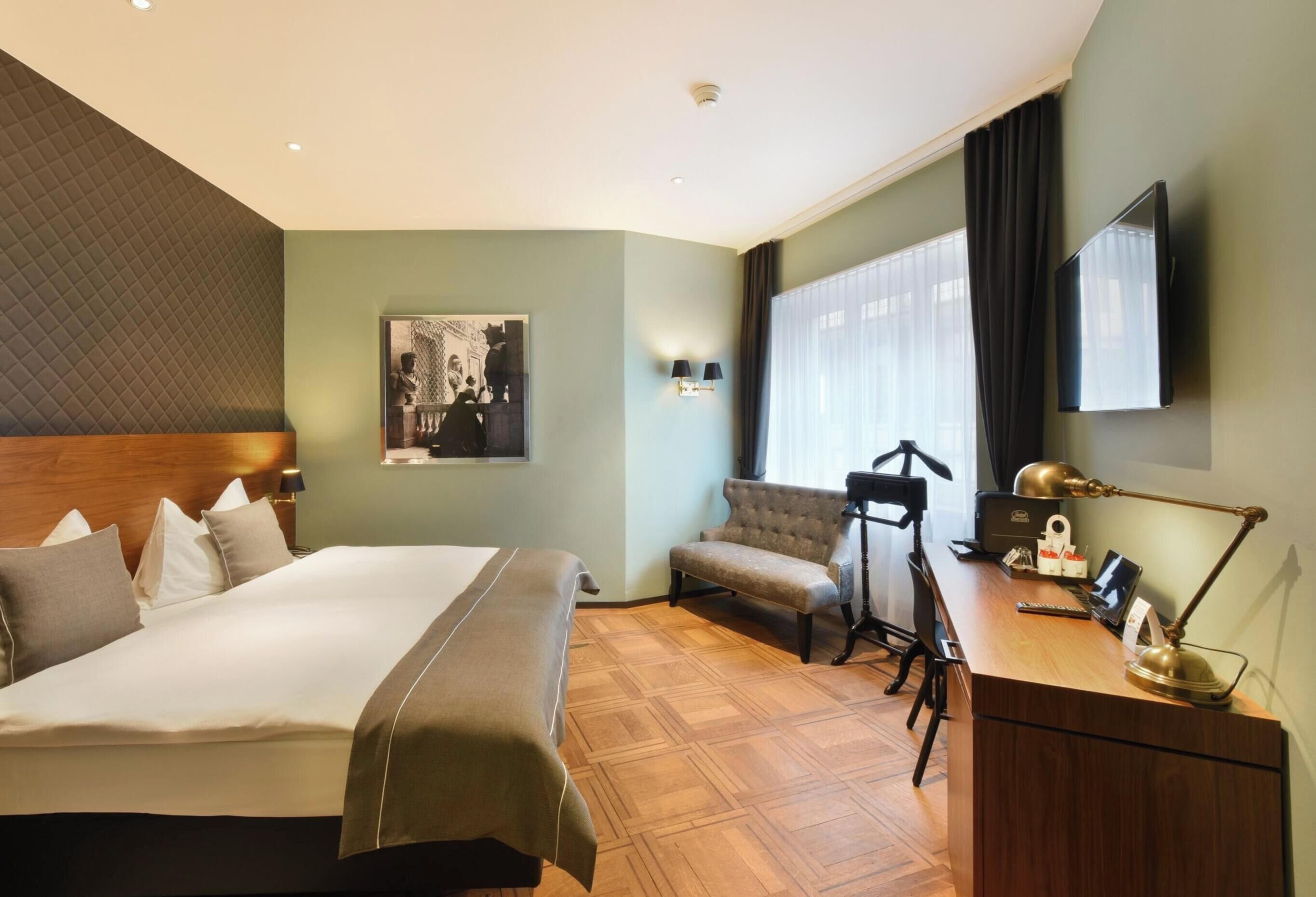 A hotel room with a bed, a desk with a lamp, a chair, a sofa, a TV, and a picture on the wall.