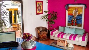 Uma sala de estar vibrante com uma parede rosa, um sofá branco com almofadas verde-água, uma cadeira de vime e um laptop sobre uma mesa azul.