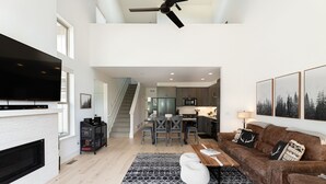 A modern living room with a fireplace, a large flat-screen TV, a brown sofa, and a dining area with a kitchen in the background.