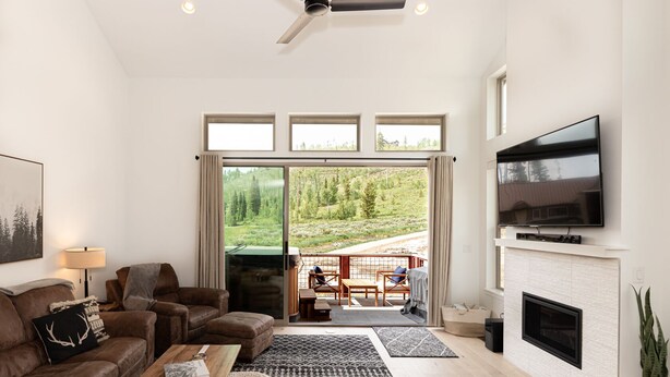 A modern living room with a sofa, coffee table, and TV. There is a fireplace and a balcony with a view of a forest.