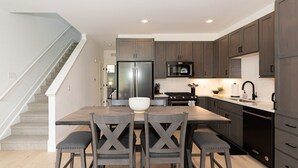 A modern kitchen with dark wooden cabinets, a stainless steel refrigerator, a microwave, and a sink. There is a wooden dining table with four chairs.