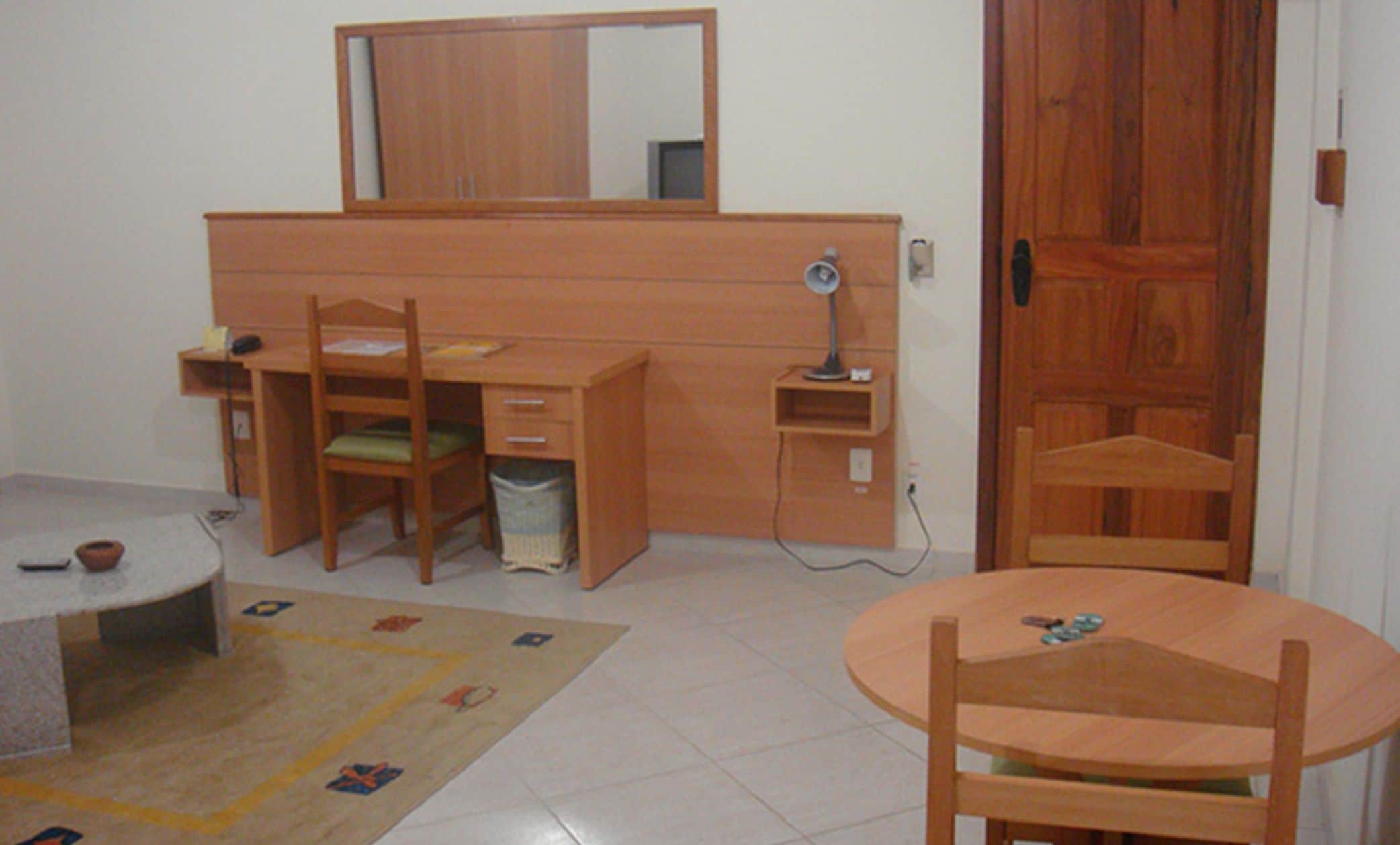 A room with a wooden desk, chair, and a small table with a chair. There is a mirror above the desk, a door to the right, and a trash bin under the desk.