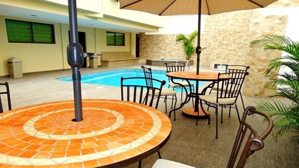 Un patio al aire libre con una mesa redonda, sillas y una sombrilla. Se observa una piscina al fondo.