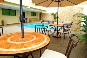 Eine Außenpatio mit einem runden Tisch, Stühlen und einem Sonnenschirm. Im Hintergrund ist ein Schwimmbecken zu sehen.