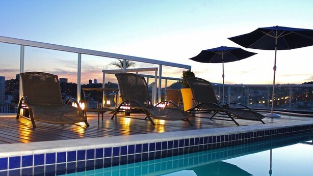 A rooftop pool area with lounge chairs and umbrellas, overlooking a cityscape at dusk. - Victory Suites (Juiz de Fora)