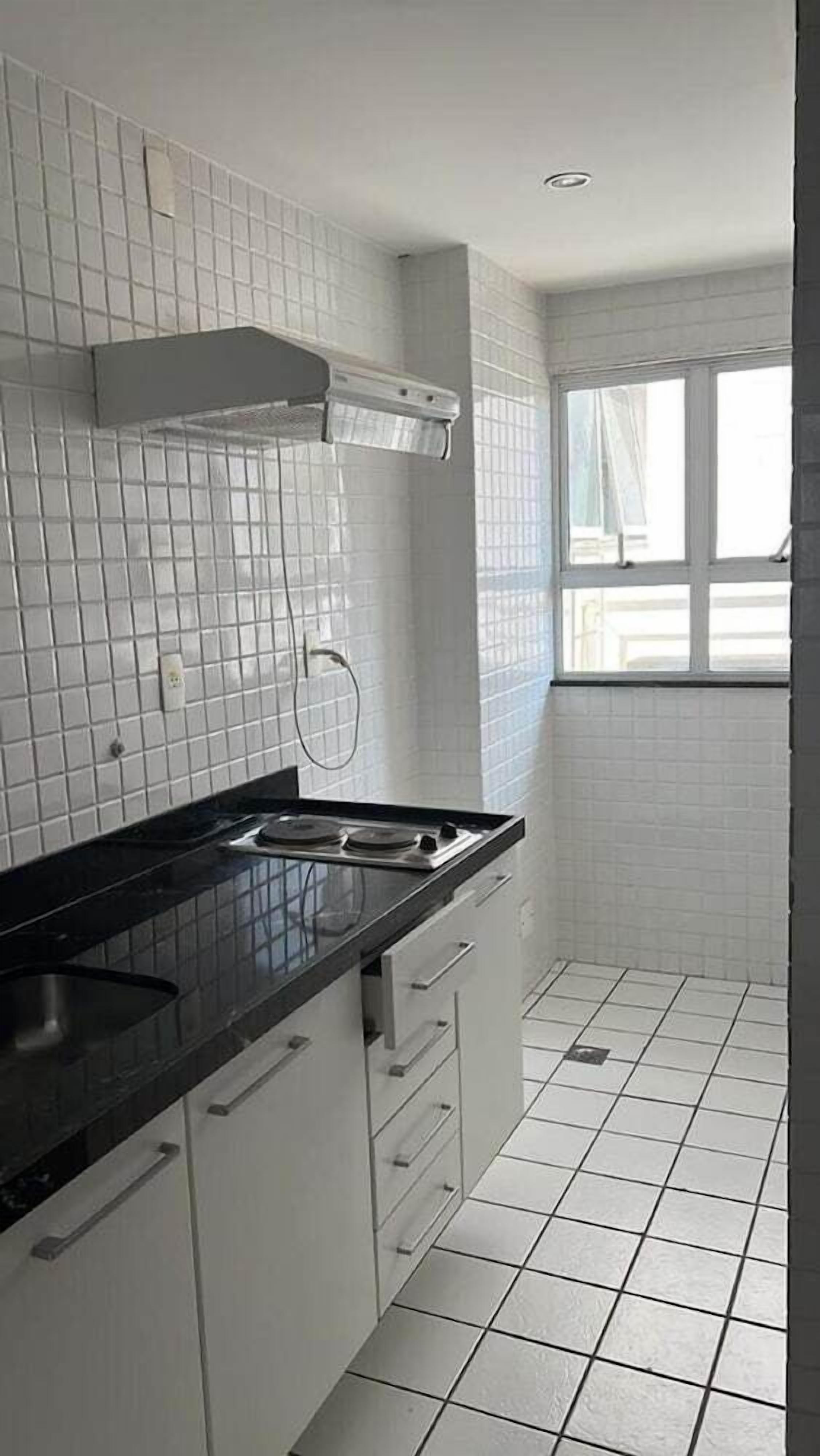 Uma cozinha com paredes de azulejos brancos, bancada preta e janela com cortinas.