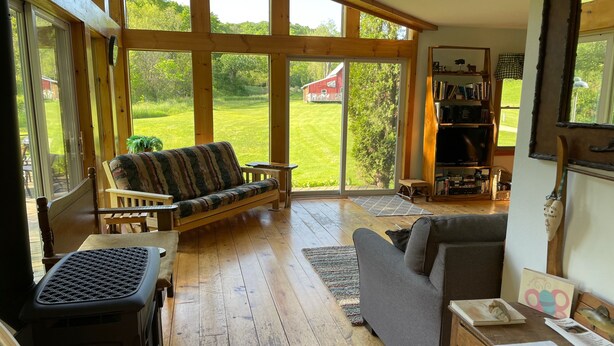 A spacious living room with large windows, a wooden floor, a sofa, a table with a fan, and a bookshelf. - Upper Place Vacation Cottage (Gays Mills)
