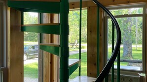 A spiral staircase with green railings and wooden steps, leading to a landing area with a window overlooking a grassy area. - Upper Place Vacation Cottage (Gays Mills)