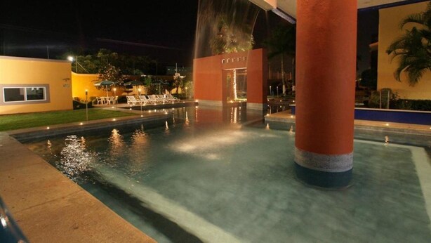 Zona de piscina nocturna con fuente, mostrador de recepción y una palmera al fondo.