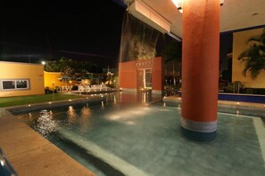 A nighttime pool area with a water feature, a reception desk, and a palm tree in the background.