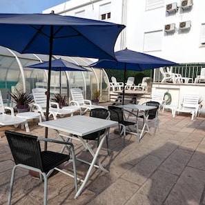 An outdoor patio with tables and chairs, covered by blue umbrellas, and white walls with air conditioning units. - Hotel Libertador Pinamar (Pinamar)