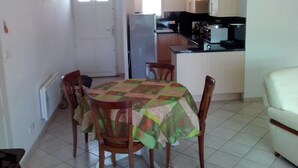 Une cuisine compacte avec des meubles blancs, un réfrigérateur et un plan de travail. On y trouve un coin repas avec une table recouverte d’une nappe à motifs floraux et des chaises en bois.