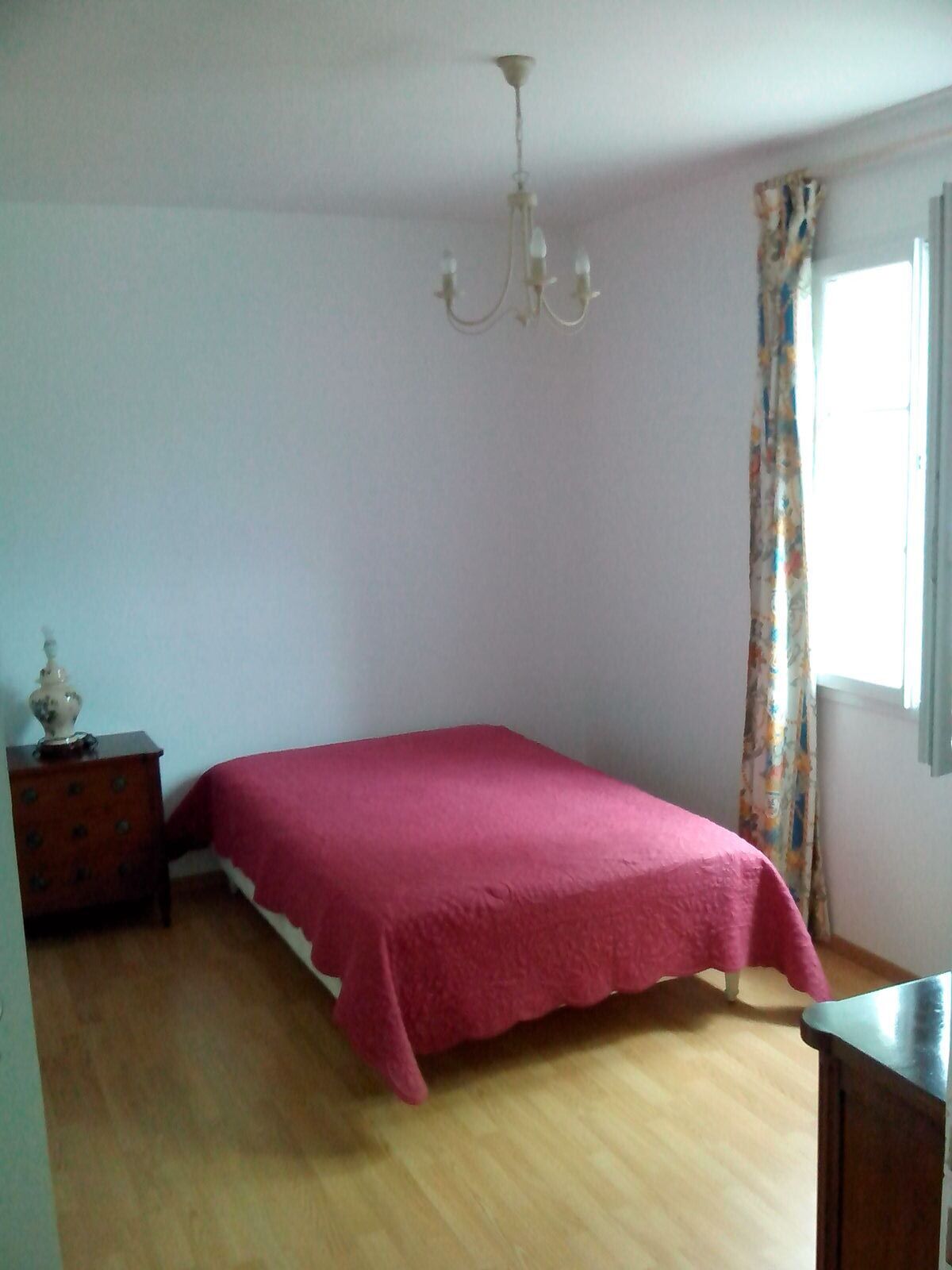 Une chambre à coucher comprenant un lit, une commode, une table de chevet, un lustre et une fenêtre avec des rideaux.
