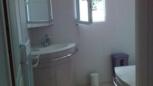 Une petite salle de bain propre, avec un lavabo blanc, un miroir et une fenêtre laissant entrer la lumière naturelle.
