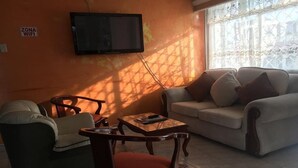 Una sala de estar con sofá, sillón y una pequeña mesa. Un televisor está montado en la pared, y un letrero indica la zona de WiFi.