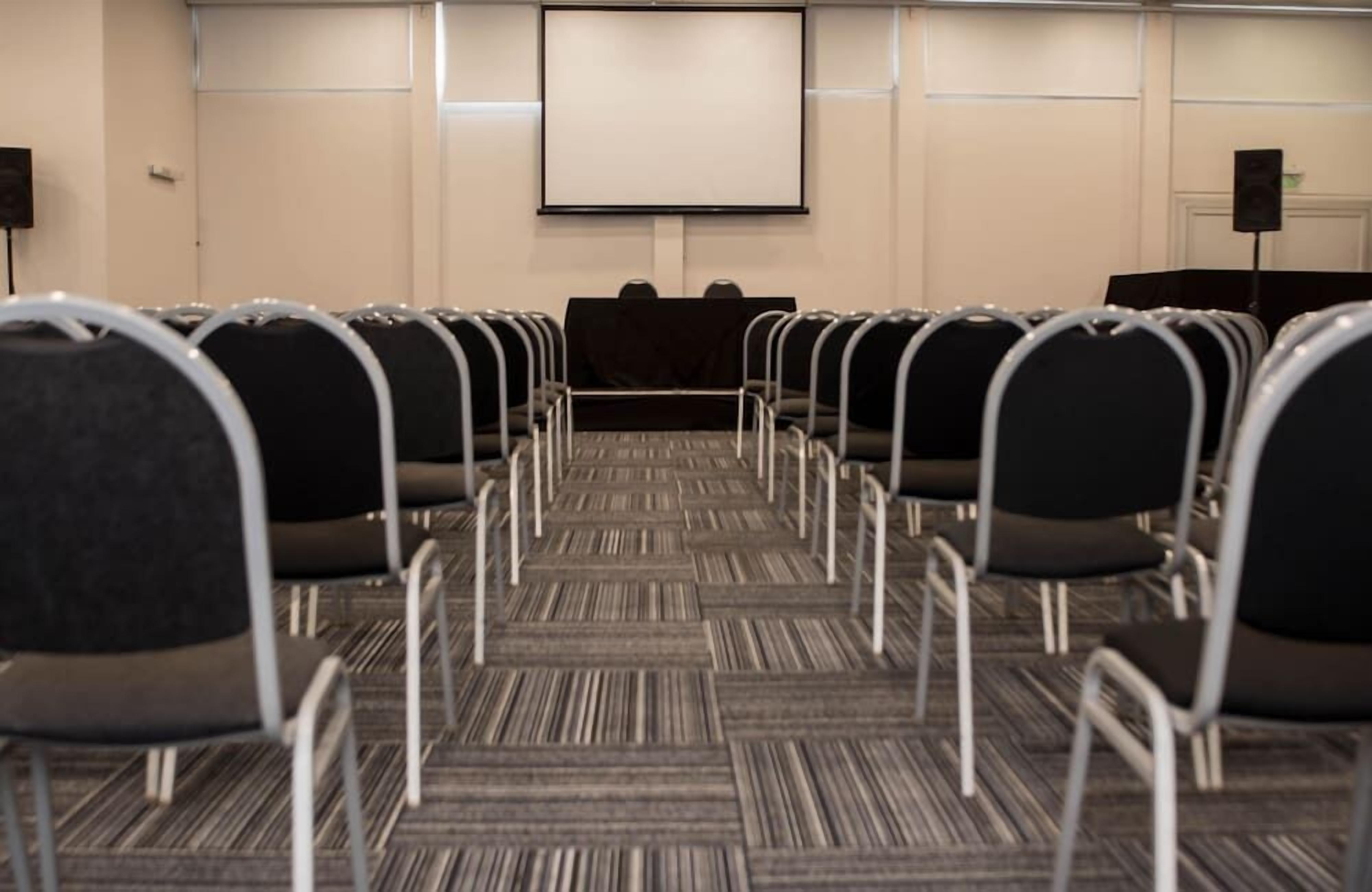 A conference room with rows of chairs facing a blank screen, carpeted floor, and a podium.
