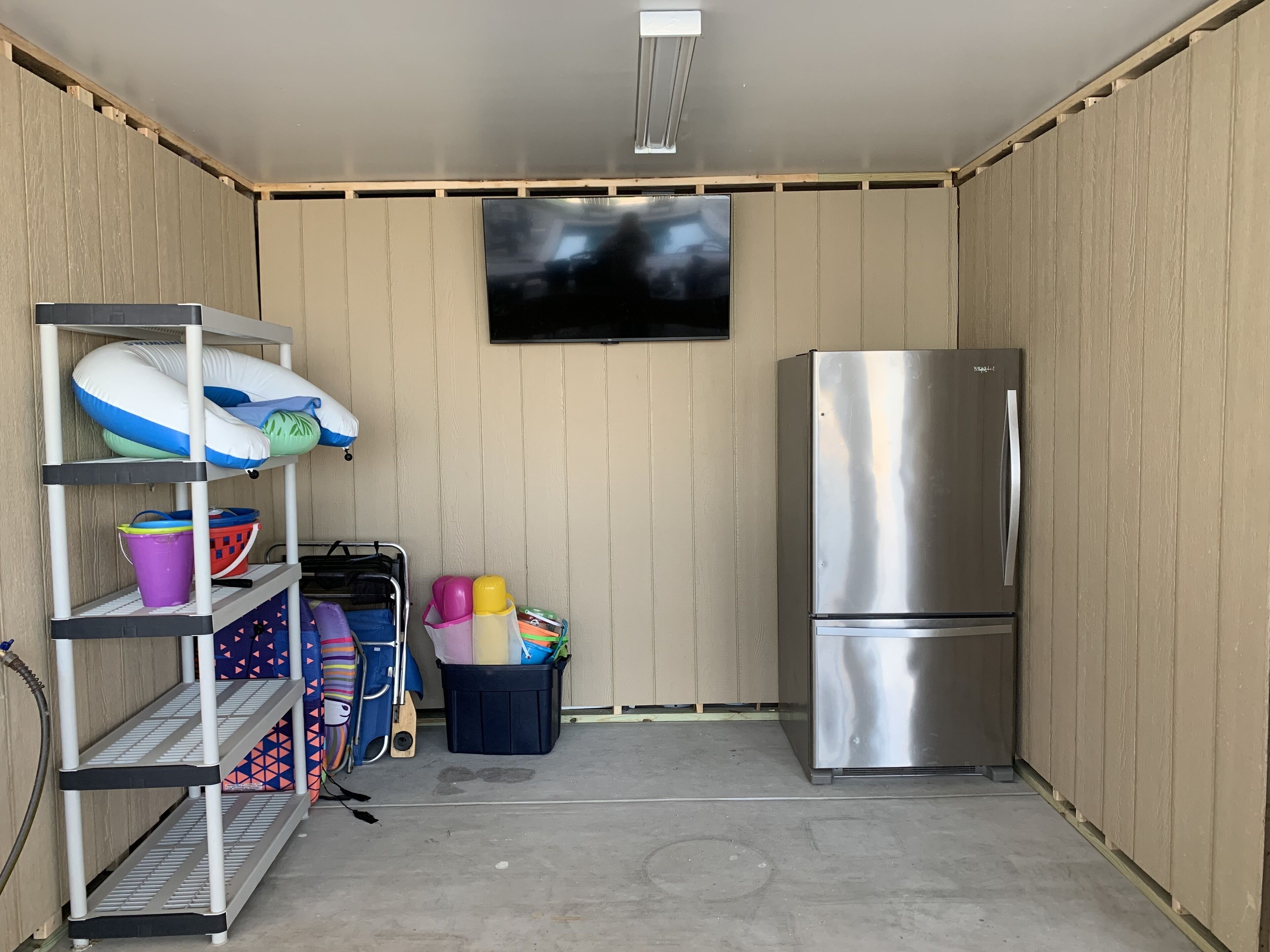 A compact kitchen with a stainless steel refrigerator, a shelf with various items, and a television mounted on the wall.
