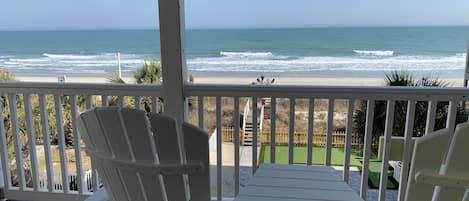 A covered deck with white chairs and a table, overlooking a beach with waves and a clear sky.