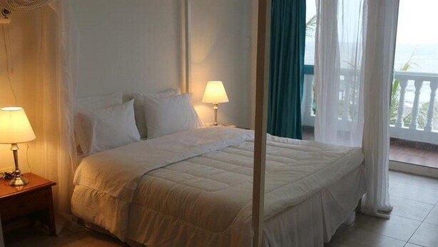 Cama bem arrumada com roupa de cama branca, duas luminárias de cabeceira e vista para o oceano através de uma varanda com cortinas brancas.