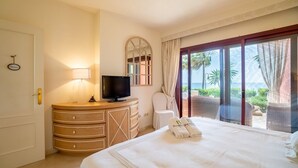 A hotel room with a bed, a TV on a wooden dresser, a bathroom mirror, and a balcony with a view of palm trees.