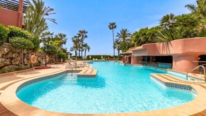 A large, clear swimming pool with a tiled edge, surrounded by palm trees and a stone wall.