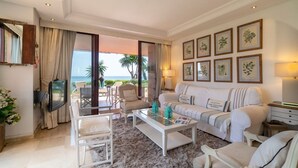 A living room with a sofa, chairs, a coffee table, and a TV. There are framed botanical prints on the wall and a view of the ocean through the glass door.
