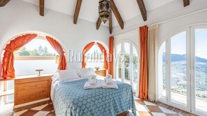 A bedroom with a bed, wooden dresser, and large windows with curtains.