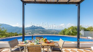 A dining area with a table set for two, overlooking a swimming pool and a mountainous landscape.