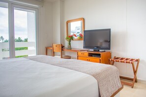 Habitación de hotel con cama, escritorio con televisor, una silla, una mesita y ventana con vista a follaje.