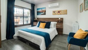 Een moderne hotelkamer met een groot bed, een bureau en een stoel. Er is een raam met gordijnen, een wandairconditioner en een kunstwerk aan de muur.