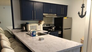 Cozinha compacta com armários azul escuro, bancada de mármore branco, fogão embutido e geladeira.