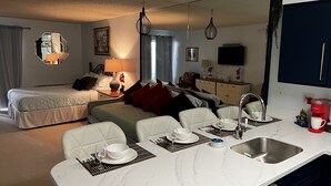 Quarto de hotel com cama, área de estar com mesa e cadeiras, cozinha com pia e máquina de café, e um espelho grande na parede.