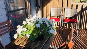 Um deck de madeira com uma mesa e cadeiras, um vaso com flores brancas e uma janela com mosquiteiro.