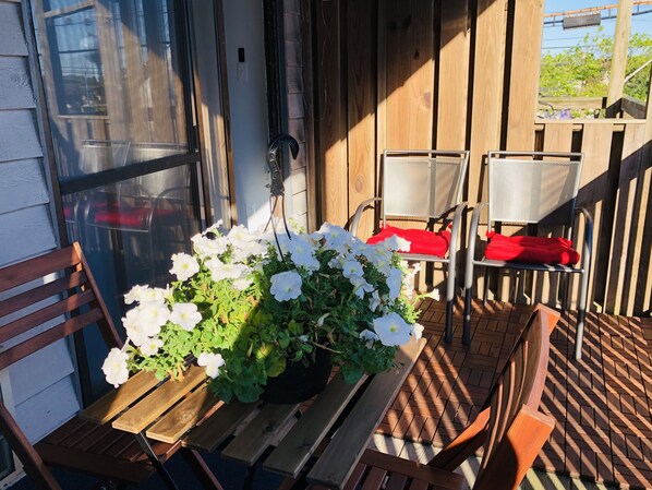 A wooden deck with a table and chairs, a potted plant with white flowers, and a window with a screen. - Newly renovated- beautiful Condo near the Beach- Sleeps 4 Linen /Towels Provided (Ocean City)
