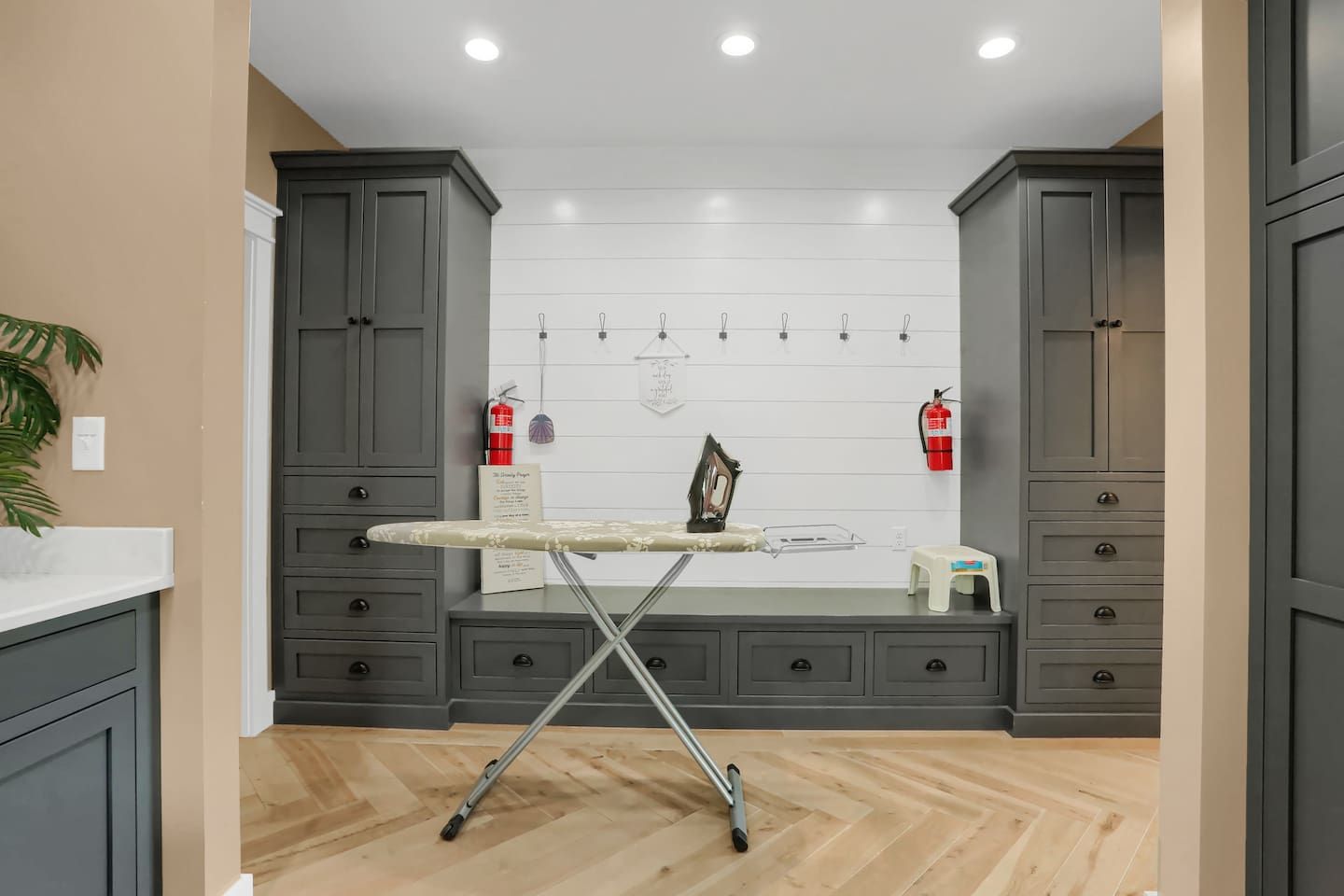 A room with a wooden floor, grey cabinets, an ironing board, and a whiteboard with hooks.