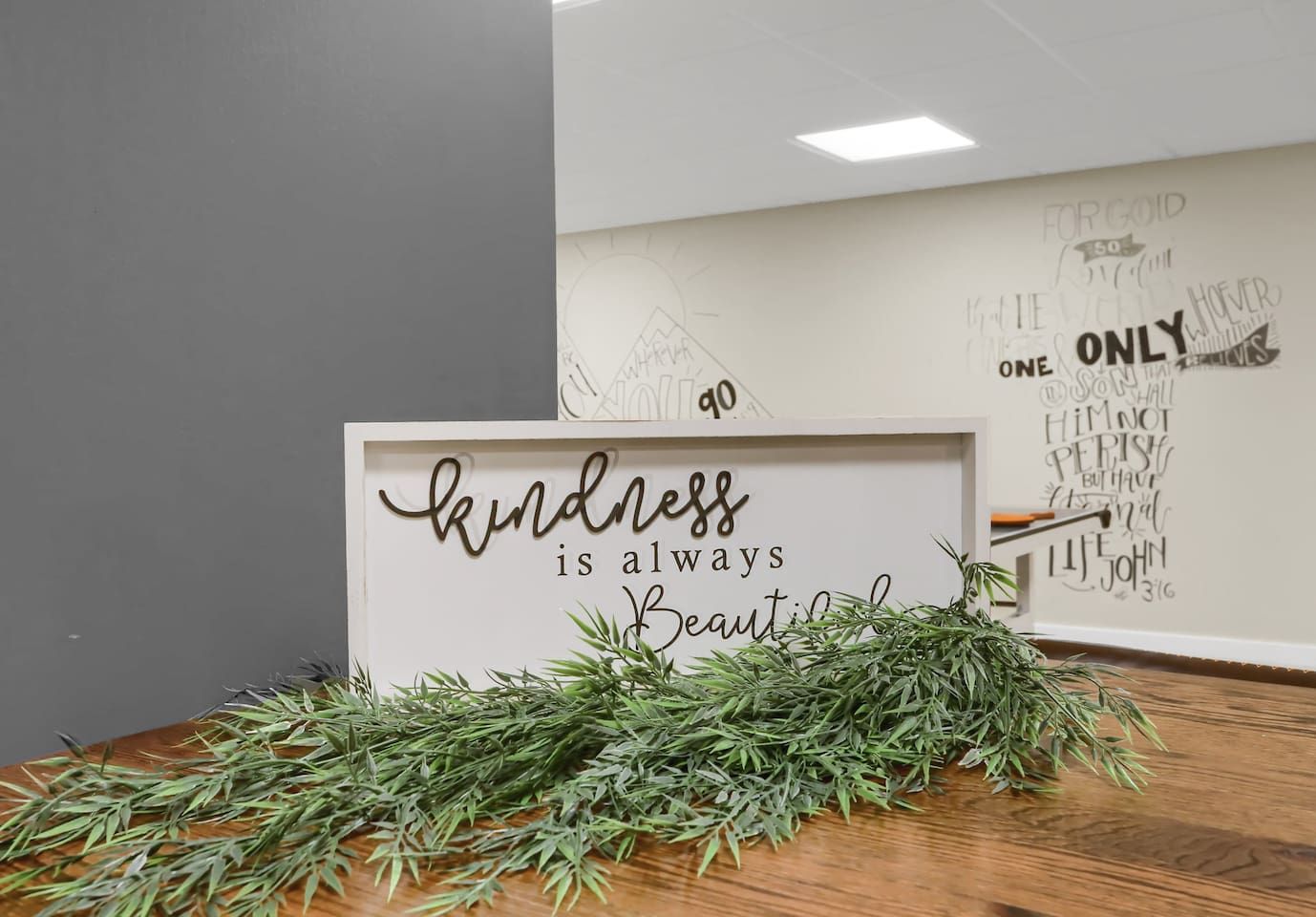 Ein gerahmtes Schild mit dem Text 'kindness is always beautiful' und ein Haufen Grünpflanzen auf einer Holztischplatte.