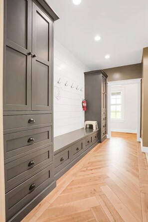 A hallway with a wooden herringbone floor, grey cabinets with drawers, a white wall with hooks, and a fire extinguisher.