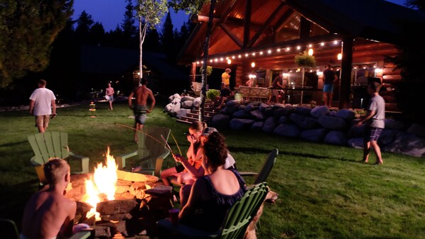 Eine Gruppe von Menschen versammelt sich um ein Lagerfeuer in einer grünen, offenen Landschaft, im Hintergrund ein Holzbauten.