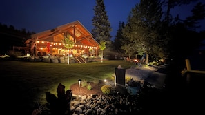 A log cabin with a porch and string lights, surrounded by a well-maintained lawn and trees. - Waterfront Log Home | Private Dock | Expansive Beautiful Yard and Gardens (Sagle)