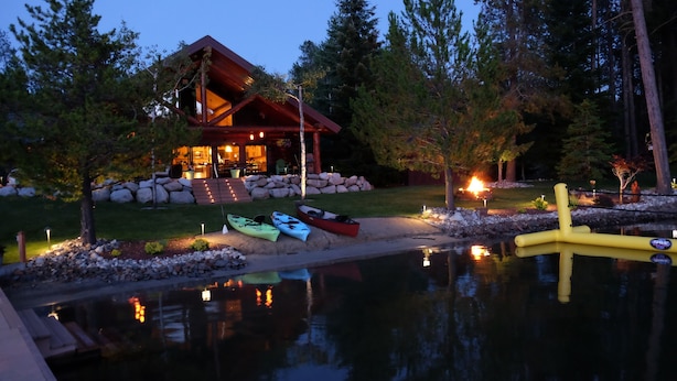 A lodge with a wooden exterior, a dock with kayaks, and a pond reflecting lights. - Waterfront Log Home | Private Dock | Expansive Beautiful Yard and Gardens (Sagle)