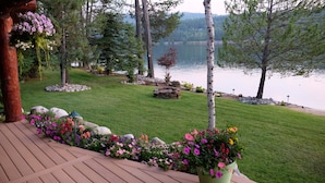 A deck with a view of a lake, surrounded by a well-maintained lawn and stone structures. - Waterfront Log Home | Private Dock | Expansive Beautiful Yard and Gardens (Sagle)