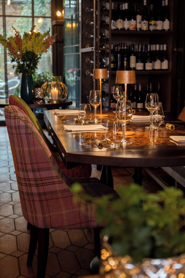Un restaurant avec une longue table dressée pour le dîner, un bar à vin en arrière-plan et une fenêtre donnant sur l’extérieur.